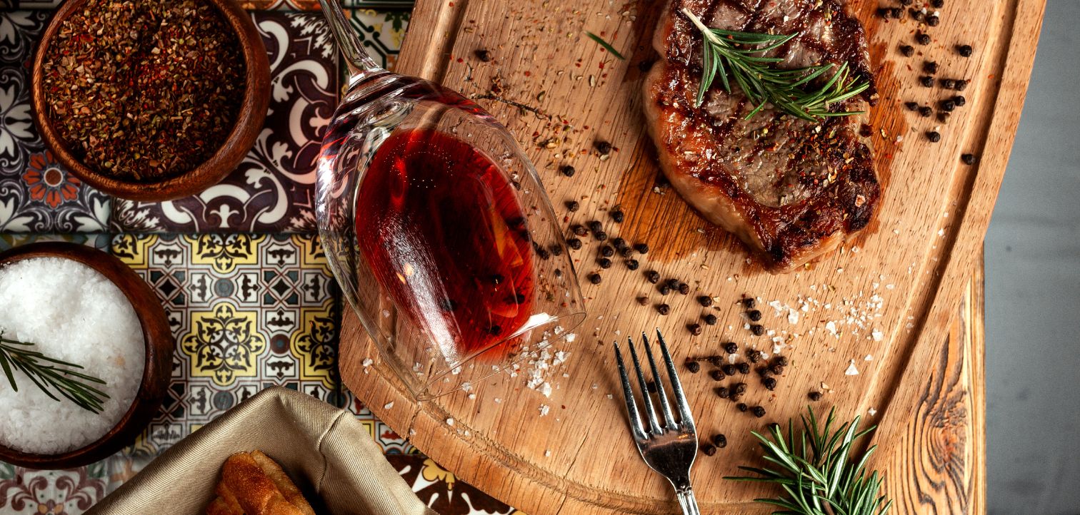 Gegrilde steak met rozemarijn op houten plank, omringd door grof zeezout, peperkorrels, een omgevallen glas rode wijn en specerijen op kleurrijke Portugese tegels.