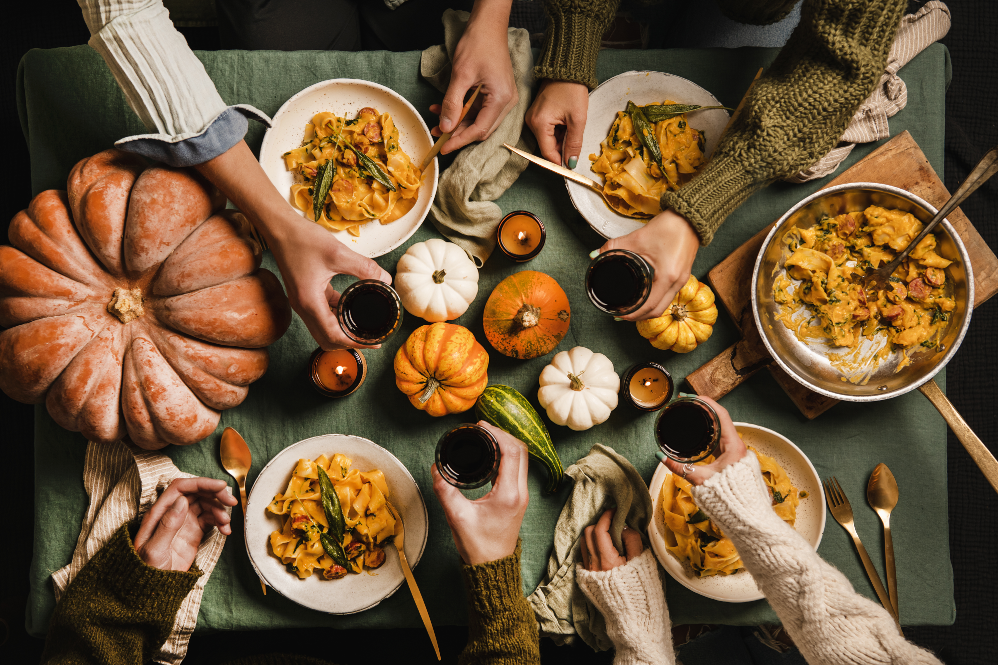 Bovenaanzicht van tafel met herfstgerechten, aangekleed met pompoenen, en vier mensen die proosten met een glas rode wijn.