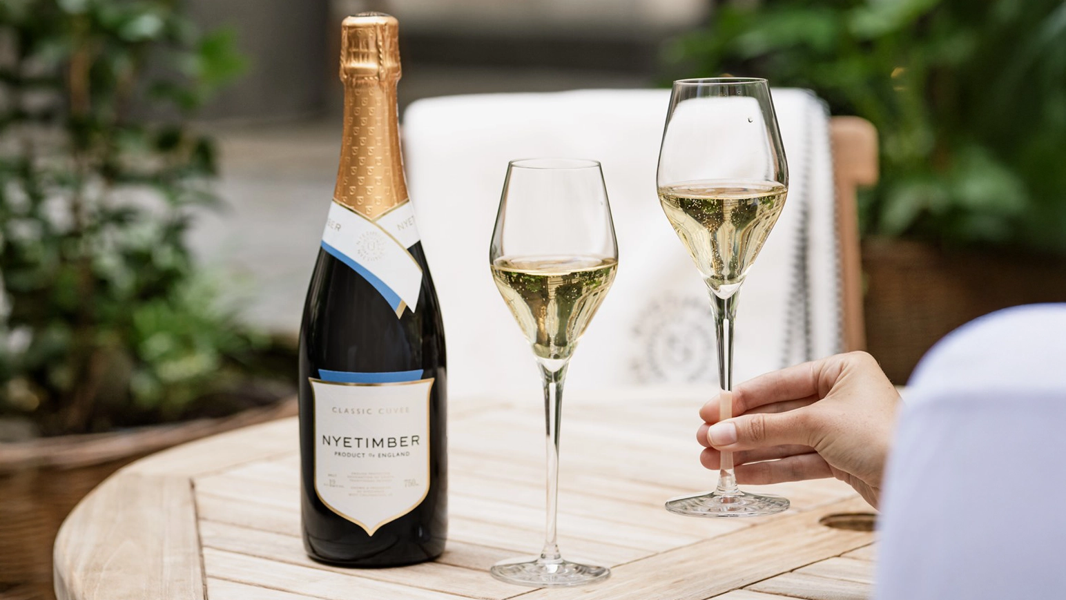 Fles Nyetimber met twee glazen mousserende wijn op een houten tafel in een zomerse tuinsetting, met een hand die een glas vasthoudt.