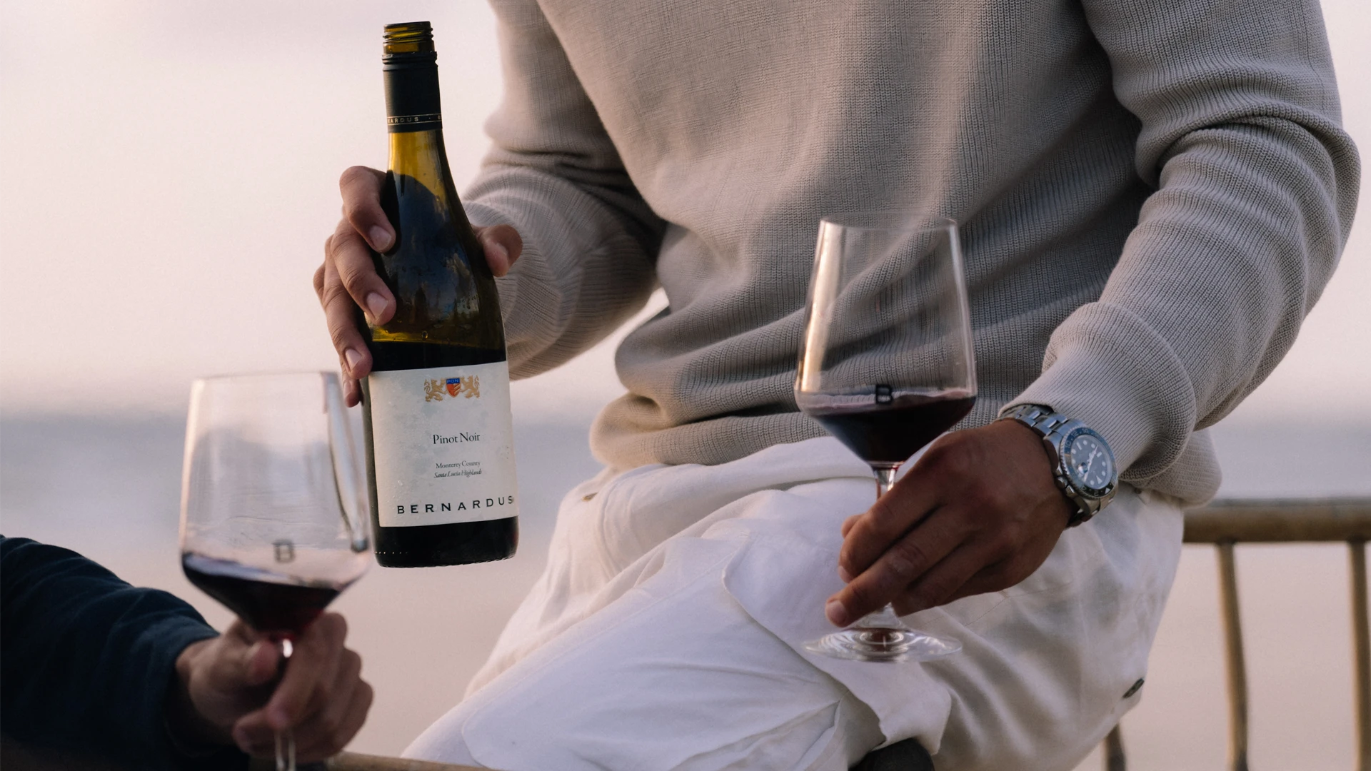 Close-up van een persoon die een fles Bernardus Pinot Noir en een glas rode wijn vasthoudt, zittend in een ontspannen setting aan zee.